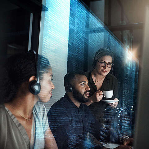 Drei Kollegen arbeiten nachts in einem Büro zusammen an einem Computer. Eine Frau mit einer Kaffeetasse blickt über die Schulter eines Mannes auf den Bildschirm, neben ihnen steht eine andere Frau mit Kopfhörern.