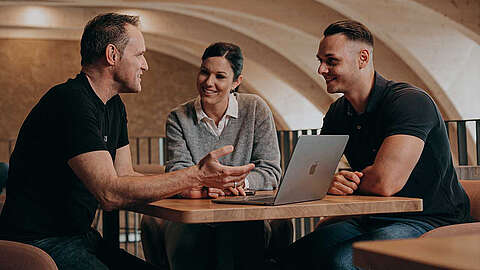 Drei Personen sitzen mit einem Laptop an einem Tisch und unterhalten sich
