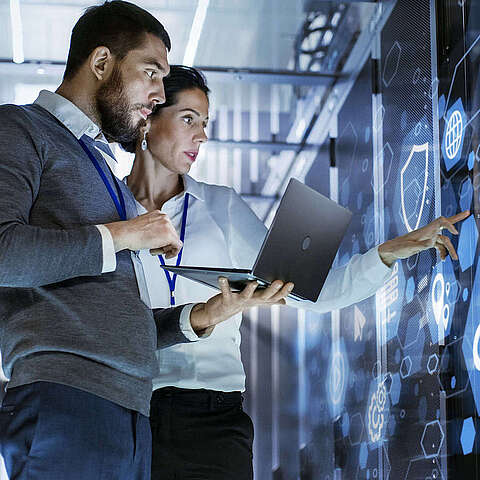 Zwei Fachleute analysieren digitale Daten auf einem transparenten Bildschirm in einem Serverraum. Einer hält einen Laptop und der andere zeigt auf den Bildschirm.