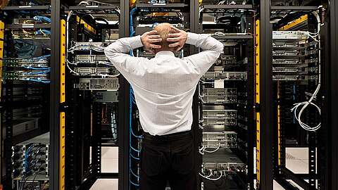 So viel Geld verschwenden Unternehmen durch heterogene Applikationslandschaften Eine Person steht mit den Händen auf dem Kopf vor einer Reihe von Server-Racks voller Netzwerkgeräte und Kabel in einem Rechenzentrum. Sie scheint die Server zu beobachten oder zu inspizieren.