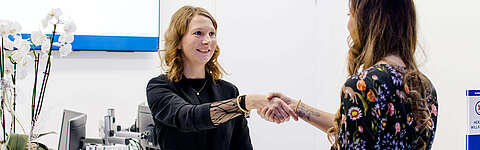 Eine Frau hinter einem Empfangstresen lächelt und schüttelt einer anderen Frau in einem geblümten Oberteil die Hand, was auf eine einladende oder professionelle Interaktion in einer Büroumgebung hindeutet.