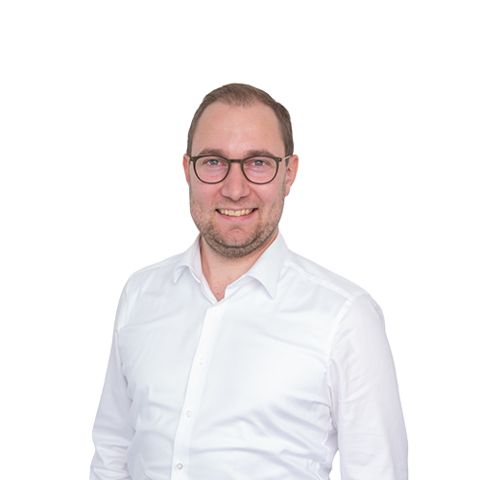 Ein Mann mit kurzen braunen Haaren und Brille, der ein weißes Button-Down-Hemd trägt, lächelt in die Kamera vor einem schlichten weißen Hintergrund.