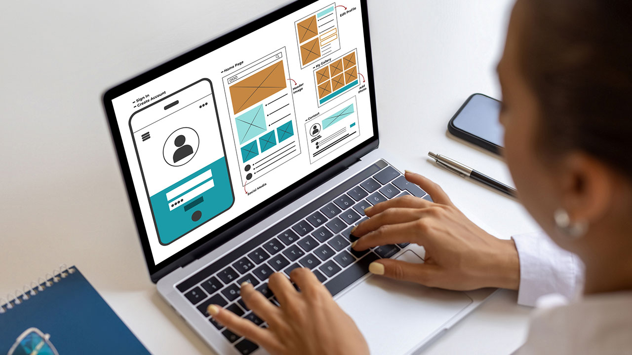 User Experience & Design Eine Person arbeitet an einem Laptop und zeigt Wireframes und Benutzeroberflächendesigns für eine Website oder App an. Ein Notizbuch, ein Stift und ein Smartphone liegen in der Nähe auf dem weißen Schreibtisch.